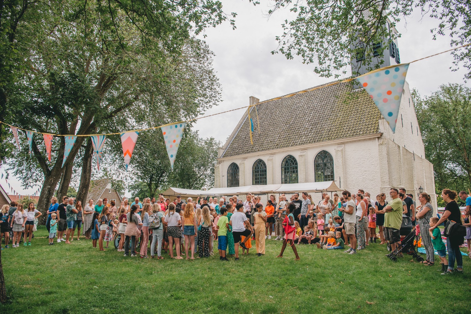 Sfeerbeeld van de zomermarkt in Groet bij het Witte Kerkje met gezellige, gekleurde vlaggetjes en een grote groep volwassenen en kinderen