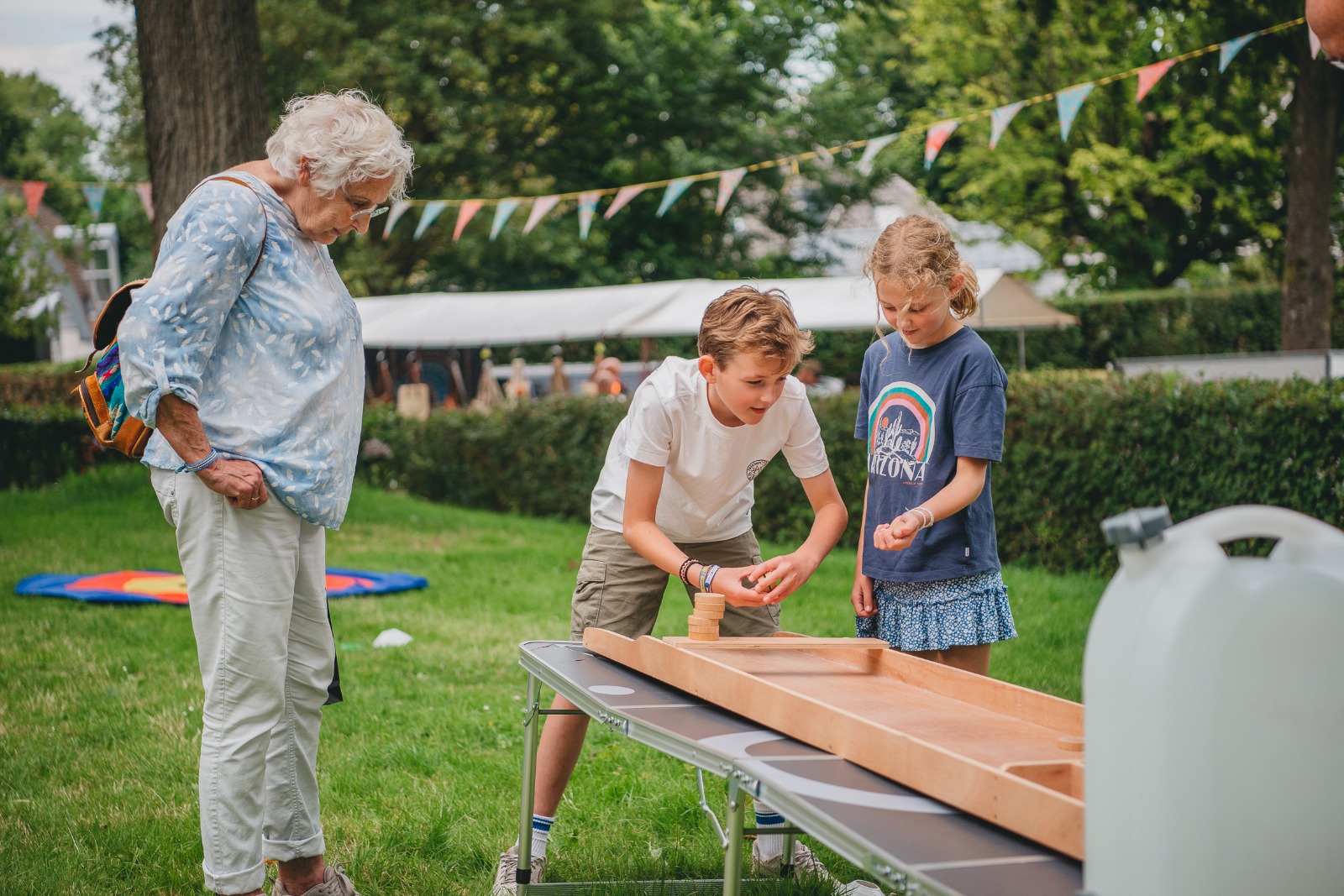 Een oma en twee kinderen die aan het sjoelen zijn op de zomermarkt met op de achtergrond vlaggetjes en de natuur van Schoorl